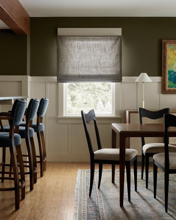 A warm, traditionally styled dining area with dark olive-green upper walls, cream wainscoting, timber flooring, and a central window fitted with a textured Roman blind. A dark timber dining table with upholstered chairs occupies the foreground, while a nearby kitchen or breakfast-bar area with tall upholstered stools is visible to the left. A slim floor lamp and framed artwork add detail to the room.