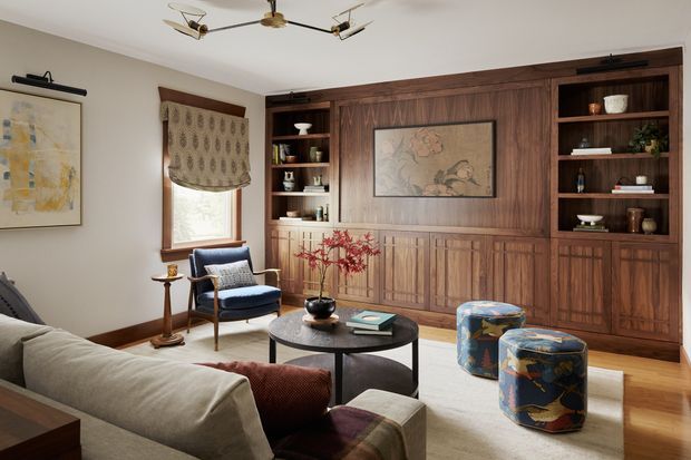 A warm, refined living room centered on a full-height dark timber built-in wall with panelled cabinetry, open display shelving, and a framed artwork niche. The room includes a light sofa, navy upholstered armchair, round black coffee table, patterned ottomans, timber flooring, a large area rug, traditional window treatment, and sculptural contemporary ceiling lighting. The overall composition blends classic timber detailing with curated modern furnishings.