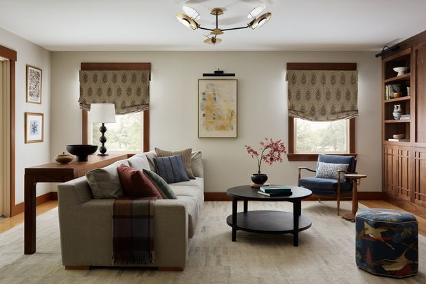 A warm, refined living room with a large neutral sofa, round black coffee table, upholstered armchair, patterned ottoman, timber console, built-in timber shelving, framed artwork, and two windows dressed with patterned Roman blinds. The room has light walls, timber trim and flooring, layered textiles, and a sculptural multi-arm ceiling light.