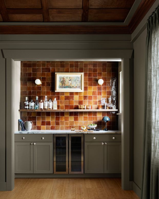 A richly detailed built-in home bar occupies a recessed alcove. Olive-grey shaker cabinetry with brass-toned knobs surrounds a central glass-front drinks refrigerator, while a dark timber shelf displays bottles and glassware. The back wall is covered in glossy, variegated terracotta-brown square tiles, illuminated by two small wall lights. A framed artwork, drinks accessories, curtains, warm timber flooring, and a dark timber coffered ceiling complete the intimate, traditional interior.