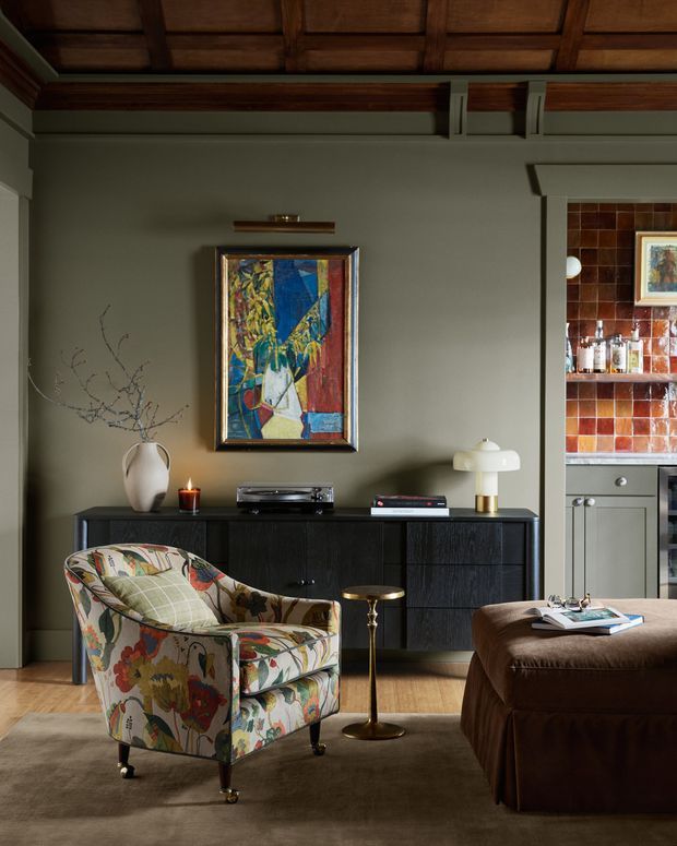 A richly styled living room with a dark wood coffered ceiling, muted green-grey walls, timber flooring, and a black built-in sideboard. A colourful patterned upholstered armchair faces a large brown ottoman, with a brass side table, ceramic lamp, artwork, record player, and decorative objects. An adjoining tiled bar area is visible through the opening on the right.