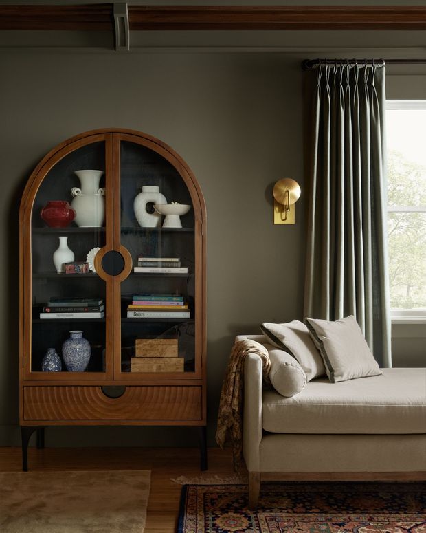A warmly lit, refined living room corner featuring a vintage-inspired arched timber display cabinet with glass doors, books and ceramic vessels, beside a cream upholstered sofa with cushions and a draped throw. Dark olive-grey walls, rich timber flooring and trim, patterned rugs, bronze-toned wall lighting, and full-length curtains create an intimate, classic atmosphere.