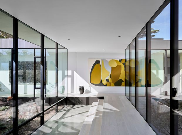 A bright, minimalist contemporary interior corridor or gallery-like living space with extensive black-framed floor-to-ceiling glazing on both sides, a pale timber or light stone floor, white walls and ceiling, and an abstract yellow, grey and black artwork at the far end. Strong sunlight casts geometric shadows across the floor, while landscaped outdoor areas and adjoining rooms are visible through the glass.