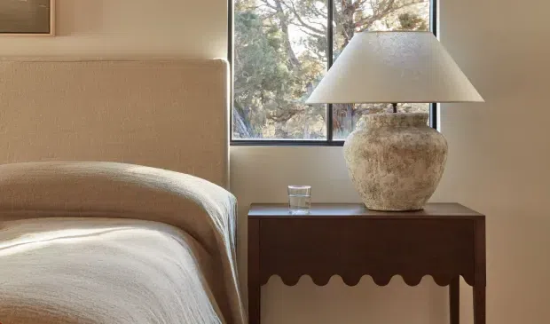 A warm, softly lit bedroom vignette featuring a partially visible upholstered bed with rounded edges, a dark wood bedside table, and a large textured ceramic lamp with a cream shade. A black-framed window overlooks leafy trees, while neutral beige and cream tones create a calm, refined atmosphere.