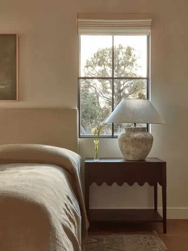 A softly lit bedroom corner with a rounded upholstered bed, warm neutral bedding, a dark wood bedside table with a scalloped apron, a textured ceramic table lamp, and a tall divided window overlooking a tree. The room has a calm, refined, earthy palette and understated decorative styling.