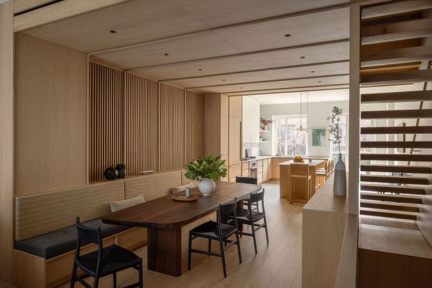 Warm, refined open-plan dining area with a dark timber dining table, built-in upholstered bench seating, black-framed chairs, and extensive light timber wall, ceiling, and cabinetry finishes. A vertical timber slat feature wall adds texture, while an open timber staircase and shelving frame the right side. The kitchen is visible beyond the dining zone, with abundant natural light and a calm, cohesive material palette.