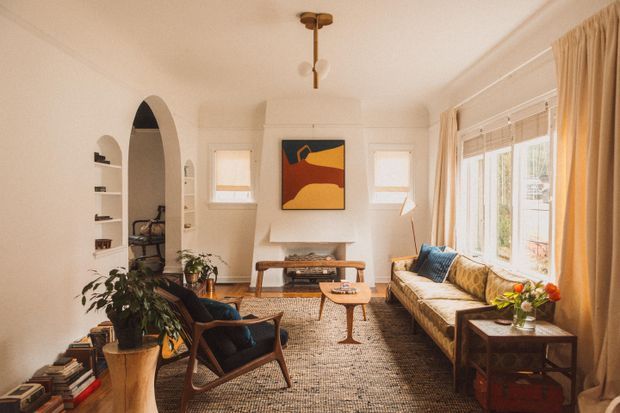 Warm, light-filled living room with a long upholstered sofa beneath large windows, mid-century-style lounge chairs, a timber coffee table, arched wall niche, fireplace, patterned rug, plants, and abstract artwork. The room features cream walls and curtains, natural timber furniture, and layered neutral textiles.