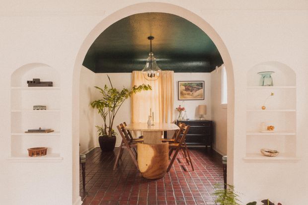 Warm, eclectic dining room viewed through a large rounded archway. The room features a dark painted ceiling, red brick tile flooring, a central wooden dining table with mixed wooden chairs, a sculptural timber chair or bench in the foreground, pendant lighting, sheer curtains, a dark sideboard, artwork, plants, and built-in arched display niches.