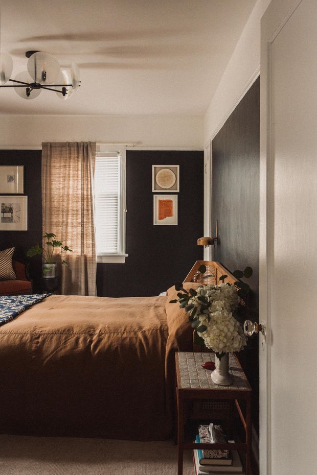 A warm, moody bedroom viewed from the doorway, featuring a dark charcoal accent wall, white ceiling and trim, a bed dressed in tan bedding, woven curtains, framed artwork, plants, and a small timber bedside table with floral arrangements. A modern sculptural ceiling light and a large dark reflective wall or mirror add visual interest.