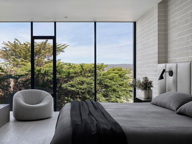 Contemporary bedroom with a low-profile upholstered bed, charcoal bedding, a sculptural grey lounge chair, and a black-framed floor-to-ceiling window overlooking dense greenery. The room features pale timber flooring, a white painted brick feature wall, minimalist black wall lighting, and a calm neutral palette.