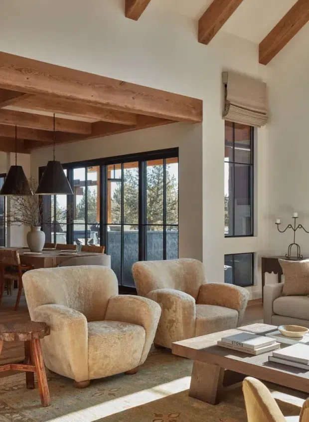 Warm, refined living and dining area with exposed timber ceiling beams, large black-framed glazed doors and windows, cream plaster walls, and plush boucle or textured beige seating. A timber coffee table, patterned rug, sculptural pendant lights, dining table, and soft Roman blinds create a sophisticated rustic interior with strong natural light.
