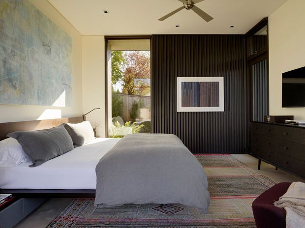 A bright, contemporary bedroom with a low-profile platform bed dressed in white and grey bedding. The room features a dark vertical slatted feature wall, matching dark built-in cabinetry and dresser, large glazing opening to a landscaped garden, recessed ceiling lights, a ceiling fan, and a layered patterned rug. Abstract artwork and a deep burgundy lounge chair add restrained colour accents.