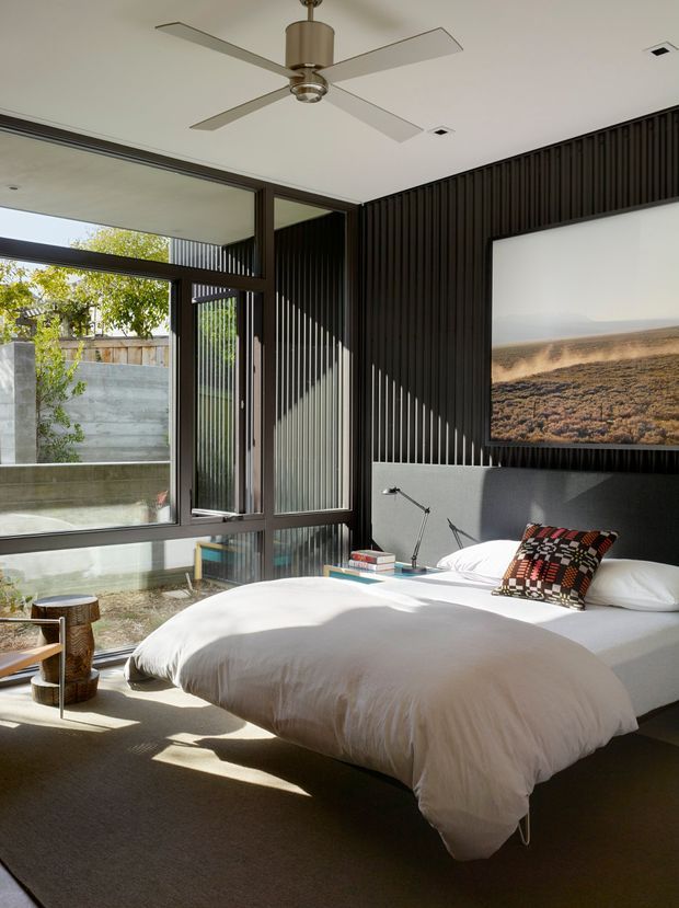 A bright contemporary bedroom with a low-profile bed dressed in white bedding, set against a dramatic dark vertically ribbed feature wall. Large floor-to-ceiling windows and glazed doors open the room to a concrete-walled outdoor area, while a ceiling fan, compact bedside lamps, a patterned accent cushion, and framed landscape artwork complete the space.