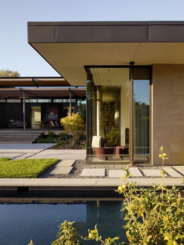 Contemporary single-storey residence viewed from a landscaped pool courtyard. The home features broad flat roof overhangs, dark metal roof-edge cladding, warm rendered walls, extensive floor-to-ceiling glazing, and a sheltered outdoor living area with timber screening. Stepping-stone paving, gravel gardens, lawn, planting, and a reflecting pool create a clean indoor-outdoor composition.