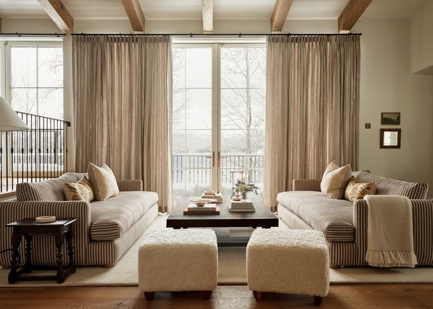Warm, refined living room with two matching striped sofas facing a dark rectangular coffee table, paired cream boucle ottomans, layered neutral cushions, and a draped throw. Floor-to-ceiling glazed doors and windows open toward a balcony, while exposed timber ceiling beams and neutral curtains add architectural character. A stair balustrade is visible at left, and framed artwork hangs on the right wall.