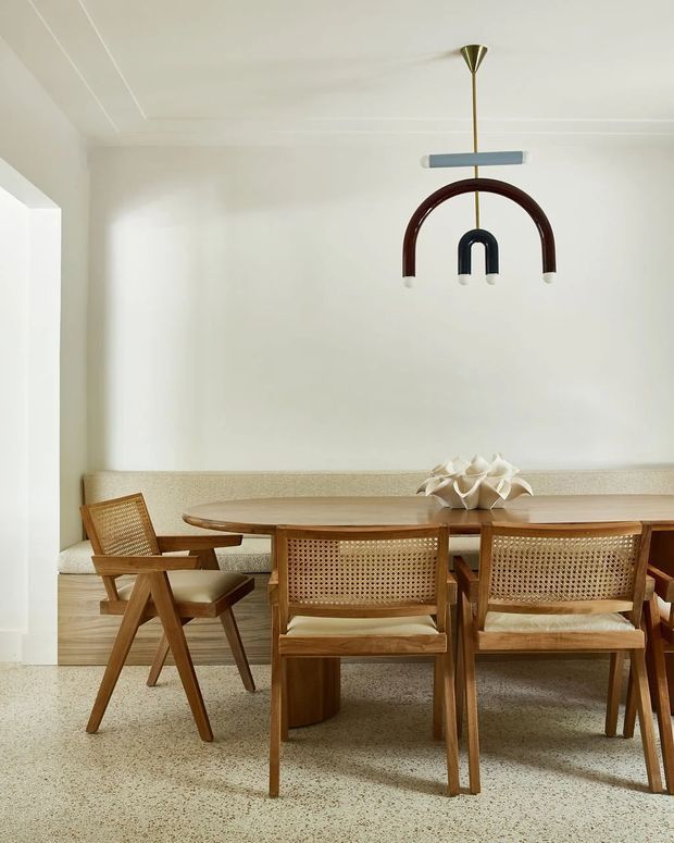 A bright, minimalist dining room with a long timber dining table, woven cane-back wooden chairs, and a built-in upholstered banquette along the wall. The space features a light terrazzo floor, white walls and ceiling with subtle linear detailing, and a sculptural mid-century-inspired pendant light with brass, dark wood, and pale blue elements. A sculptural floral-like centerpiece sits on the table.