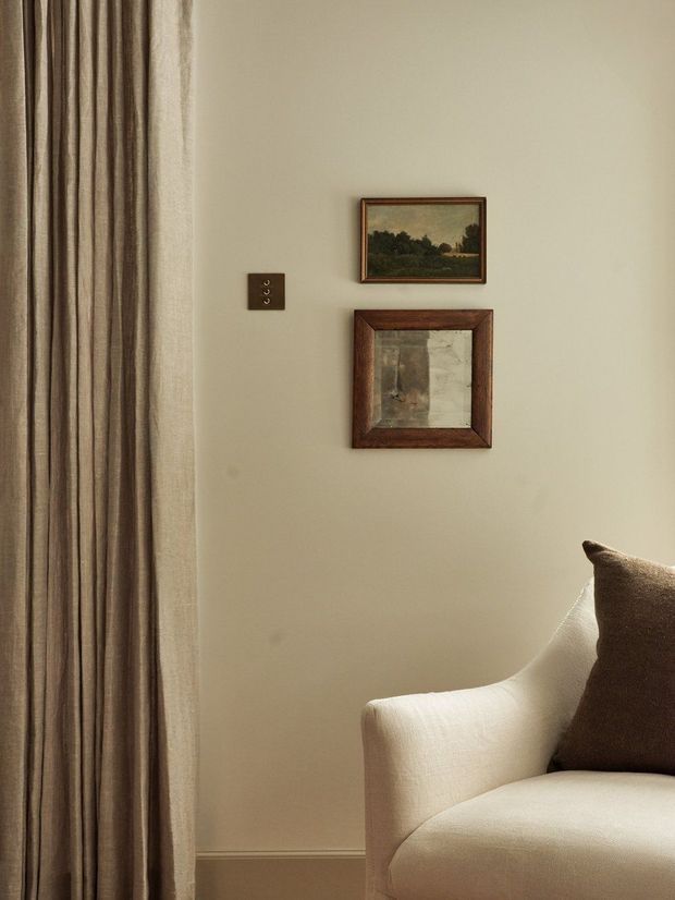 A softly lit interior detail showing a neutral beige wall with two small framed artworks, a dark wall switch plate, full-height taupe curtains, and the corner of a cream upholstered armchair with a brown cushion. The composition has a warm, traditional and understated character.