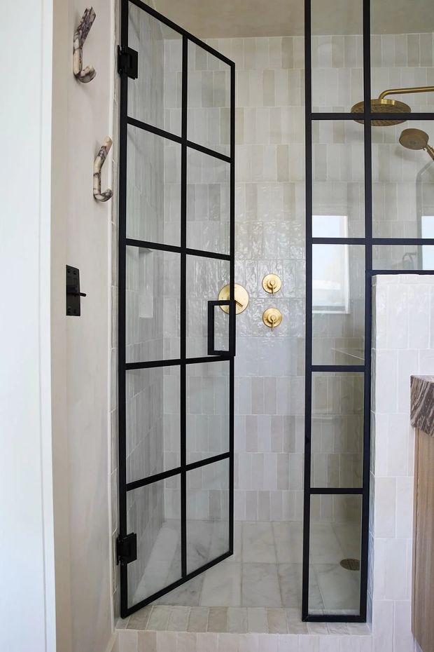 A bright contemporary bathroom shower enclosure with a black metal-framed glass door and fixed glass panel. The shower walls and threshold are clad in pale textured rectangular tiles, with white marble-look floor tiles, brass/gold shower controls and fixtures, and a partial vanity or partition visible at right.
