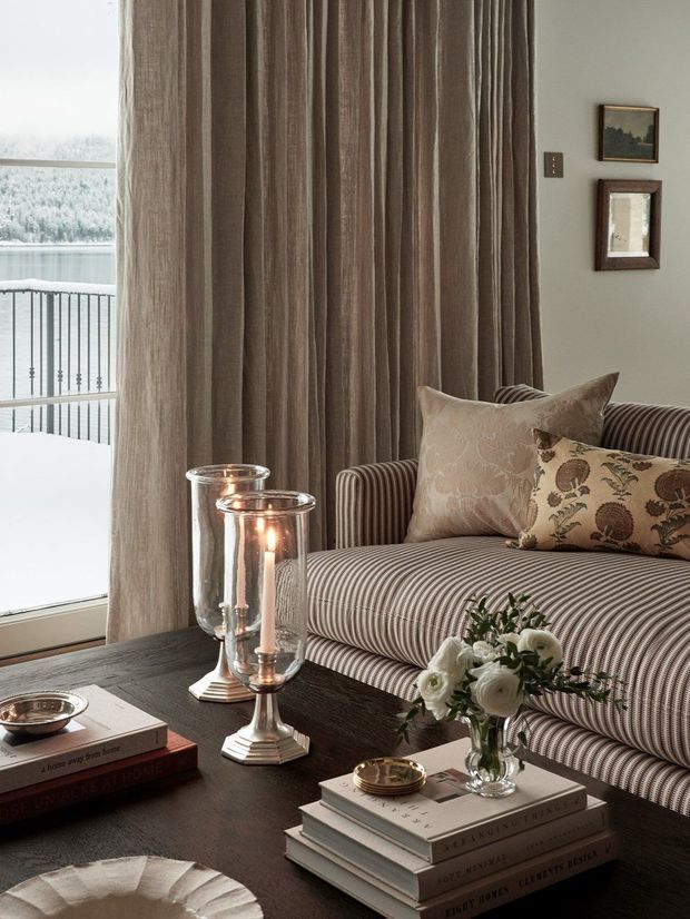 Elegant sitting area with a striped upholstered sofa, patterned decorative cushions, floor-to-ceiling taupe curtains, framed artwork, and a dark coffee table styled with books, candles in glass hurricanes, a small dish, and a floral arrangement. Large glazing at left overlooks a snowy wooded landscape and balcony railing. The room has a refined, traditional feel with warm layered neutrals and soft natural light.
