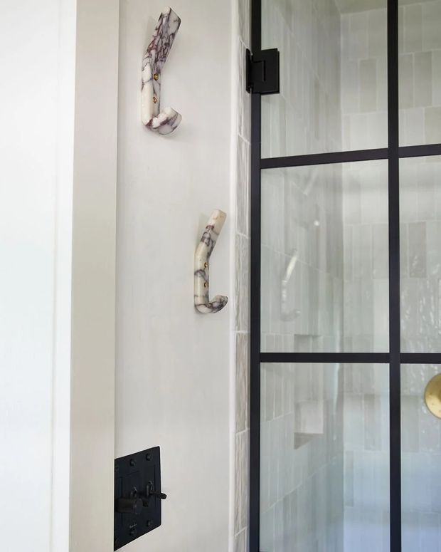 Close-up of a bathroom shower area featuring a black-framed glass shower screen, pale tiled walls, matte black shower controls, and decorative sculptural wall hooks. The image has a bright, refined contemporary look with subtle industrial detailing.