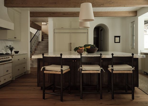A refined traditional kitchen with a large central island and three woven-seat bar stools. The room features dark timber flooring, dark timber island cabinetry, pale painted cabinetry, a light stone benchtop, exposed timber ceiling beams, pendant lighting, and a stairway leading to an adjacent level.