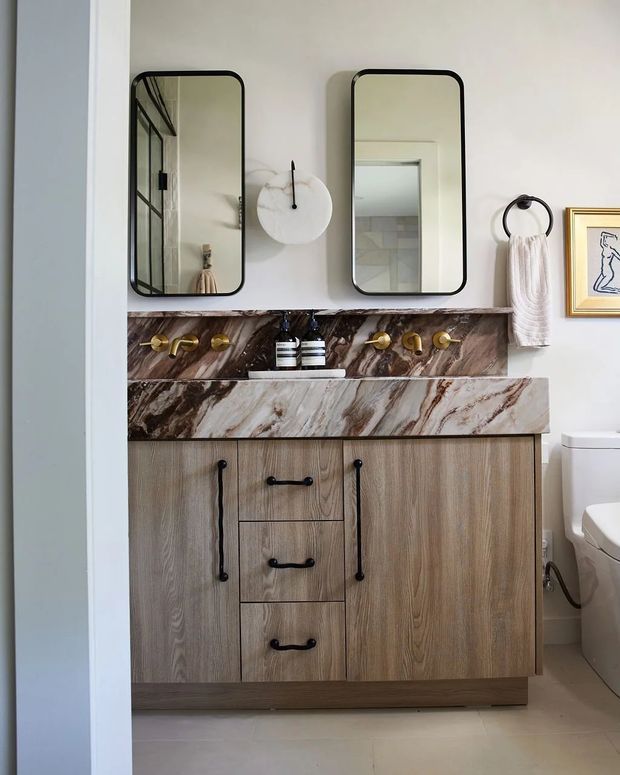 A compact, refined bathroom vanity area with a double-mirror arrangement, wall-mounted brass plumbing fixtures, a dramatic veined stone backsplash and countertop, and a warm wood-grain vanity with black pulls. A toilet is visible at right, along with a towel ring and framed artwork. The space combines natural stone, timber-look joinery, aged brass, and soft neutral finishes.