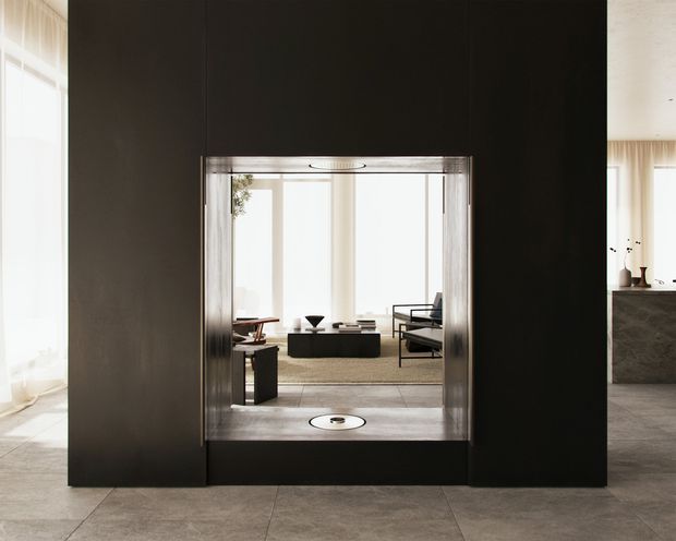 Interior view facing a compact residential elevator set within a dark feature wall, with a bright contemporary living room visible beyond. The space has large pale floor tiles, sheer curtains, strong natural light, dark wall surfaces, and restrained modern furniture. A partial kitchen or stone-clad counter is visible at right.