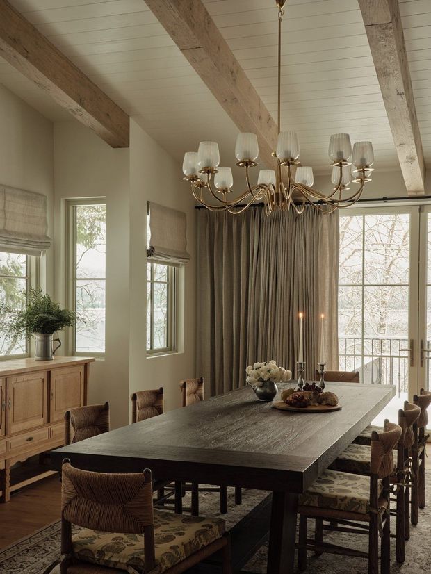 Warm, refined dining room with a large rustic timber dining table surrounded by upholstered woven chairs. The room features exposed chunky timber ceiling beams, a white painted plank ceiling, tall windows with Roman shades and floor-length drapery, built-in timber cabinetry, and an ornate brass multi-arm chandelier. Neutral taupe, cream, and natural timber tones create an inviting traditional country-inspired interior.
