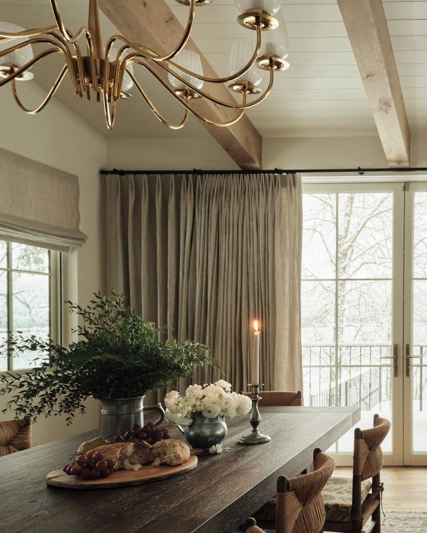 A refined dining area with a long dark timber dining table, woven upholstered dining chairs, layered neutral curtains, floral arrangements, and candlelight. The room features a pale painted timber-plank ceiling with exposed timber beams, large glazed doors and windows, and an ornate brass sculptural chandelier. The overall scheme is warm, traditional, and understatedly luxurious.