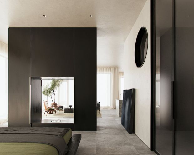 A refined contemporary interior view from a bedroom toward an adjoining living area. The foreground shows part of a bed with layered neutral bedding, while a tall dark built-in wall and mirrored opening frame a bright sitting space beyond. Pale textured walls, large-format grey floor tiles, sheer curtains, recessed ceiling lights, dark-framed glazing or cabinetry, and a round black wall feature create a calm, minimalist composition.