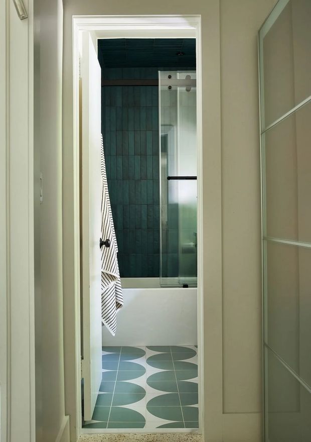 View through a doorway into a compact bathroom featuring a white bathtub with clear glass shower screen, dark teal vertical wall tiles, and a geometric teal-and-white tiled floor. The surrounding hallway has pale green walls, white trim, and a large framed mirror or glazed panel.
