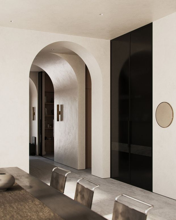 A refined contemporary dining area with a long dark stone dining table and sculptural metal-framed chairs in the foreground. The room features warm off-white textured plaster walls, a large rounded archway leading to a darker adjoining space, and tall glossy black built-in cabinetry. Minimal recessed ceiling lights, dark timber accents, and a small round wall mirror create a restrained, luxurious interior.