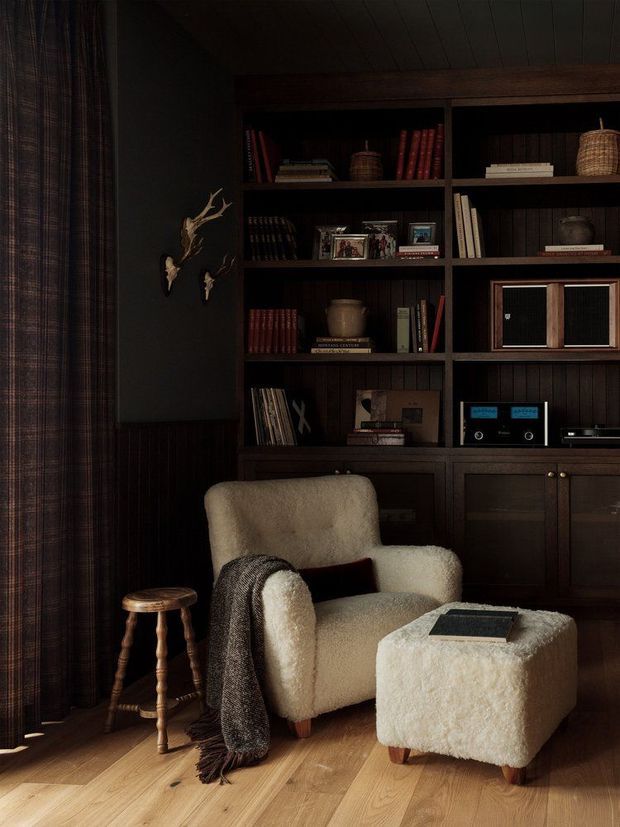 A moody, intimate reading nook with a cream boucle armchair and matching ottoman positioned in front of dark built-in timber shelving. The shelving is filled with books, framed photographs, baskets, a vintage-style audio system and decorative objects. Dark wood wall panelling and cabinetry contrast with light timber flooring, while a small turned timber stool, draped throw and antler wall ornament add warmth and character. Floor-length dark plaid curtains frame the left side.