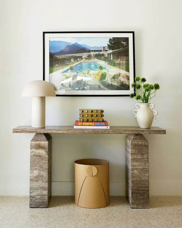 A styled hallway or entryway vignette with a stone console table against a pale wall. The console has chunky stone supports and a textured stone top, with a sculptural cream table lamp, stacked books, a small gold-toned decorative object, and a white ceramic vase with green stems. A black-framed artwork hangs above, and a sculptural tan wastebasket sits beneath the console on a terrazzo-like floor.