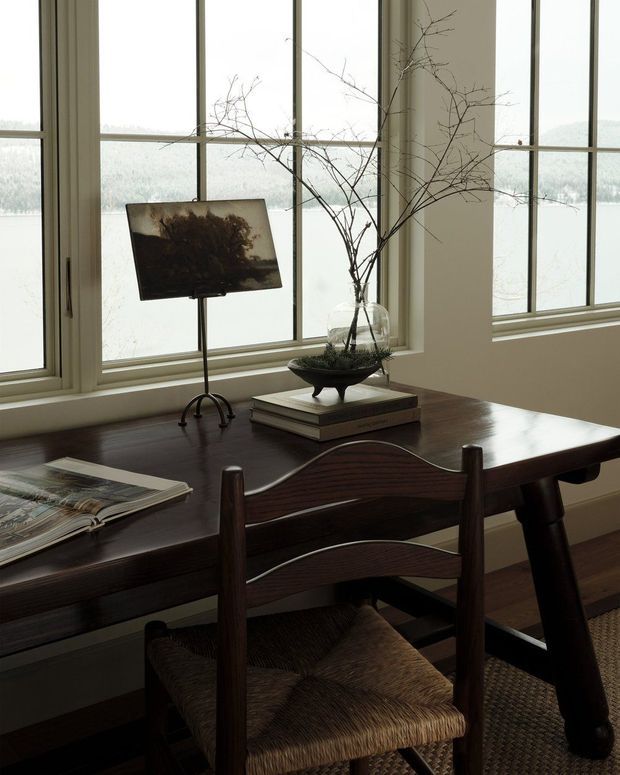A warmly lit study or workspace positioned beside large multi-pane windows overlooking a snowy landscape. The room features a dark timber desk, a woven-seat wooden chair, a small table lamp, stacked books, a glass vase with bare branches, and a textured floor covering. The restrained natural palette and vintage furnishings create a quiet, refined atmosphere.