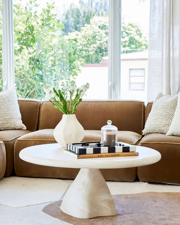 Bright, inviting living room vignette with a low caramel-brown sectional sofa positioned in front of large windows overlooking leafy greenery. A sculptural off-white round coffee table with a pedestal base holds a ceramic vase, books, and a glass cloche. Textured cream cushions, sheer curtains, a pale rug, and natural light create a warm contemporary interior.