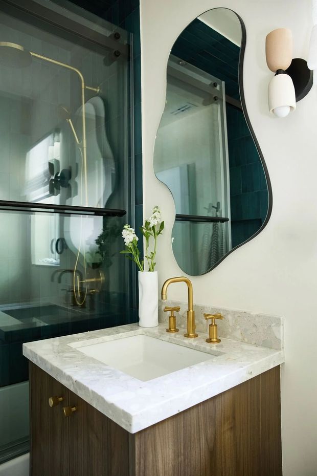 A refined bathroom vanity area featuring a dark timber-grain cabinet, pale natural stone benchtop and integrated white basin. Brushed gold tapware contrasts with deep teal tiled shower walls and black shower hardware. A sculptural organic-shaped mirror, contemporary wall sconce, and white floral arrangement add decorative character. The image is photographed from within the bathroom, with the glass-enclosed shower visible beside the vanity.