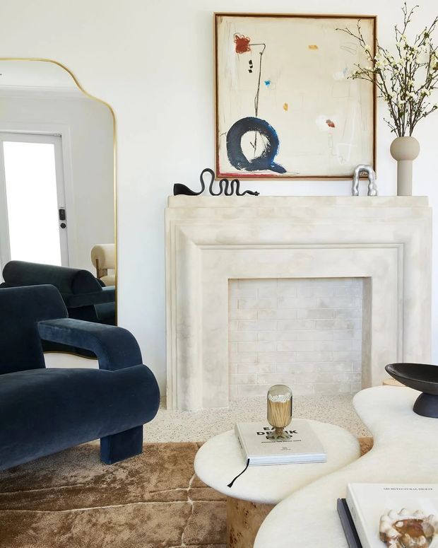 A refined living room vignette centred on a pale stone or plaster fireplace surround with a white brick-lined firebox. The room features sculptural deep navy upholstered lounge chairs, rounded cream coffee tables, a textured neutral rug, a large organic abstract artwork in a timber frame, an oversized arched mirror, and a ceramic vase with branches. The overall composition is light, artistic, and softly luxurious with rounded contemporary forms.
