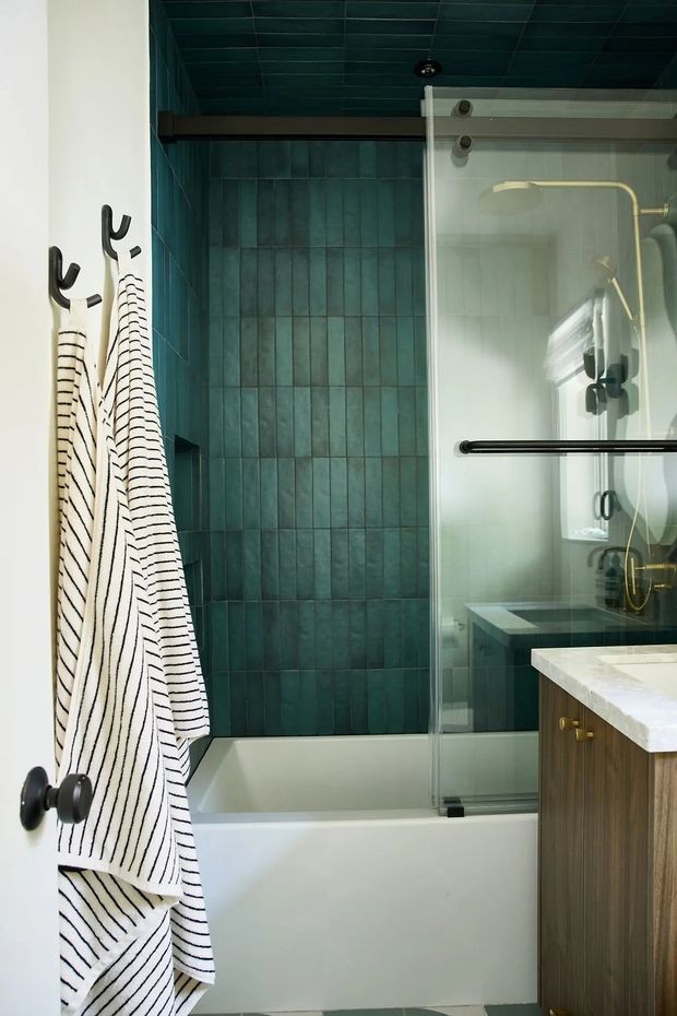 Compact bathroom with a white bathtub and glass shower screen, clad in dark teal-green rectangular wall and ceiling tiles. A timber vanity with a pale stone benchtop and brass hardware occupies the right side, while striped towels hang from black wall hooks. The space has a moody, refined contemporary feel with black and brass fixtures.