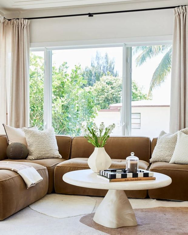 Bright, inviting living room with a large tan leather sectional sofa arranged around a sculptural white oval coffee table. The room features floor-to-ceiling windows with cream curtains, abundant greenery visible outside, neutral textured cushions, a pale area rug, and a simple vase with greenery on the table.