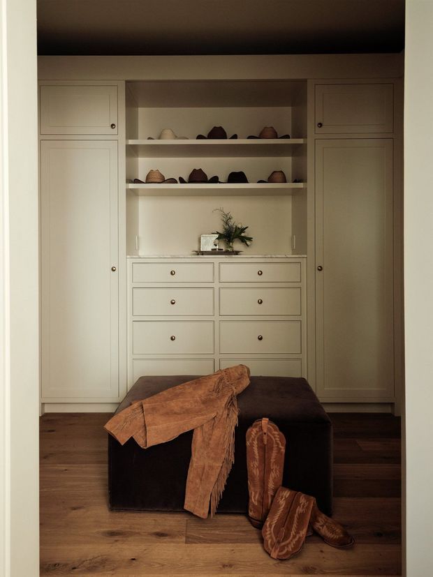 A warm, carefully fitted dressing and storage area viewed through a doorway. The room features full-height painted cabinetry with paneled doors, a central bank of drawers, open shelves displaying hats, and a dark upholstered ottoman in front with a leather garment and boots. Dark timber flooring and soft warm lighting create an intimate, refined atmosphere.