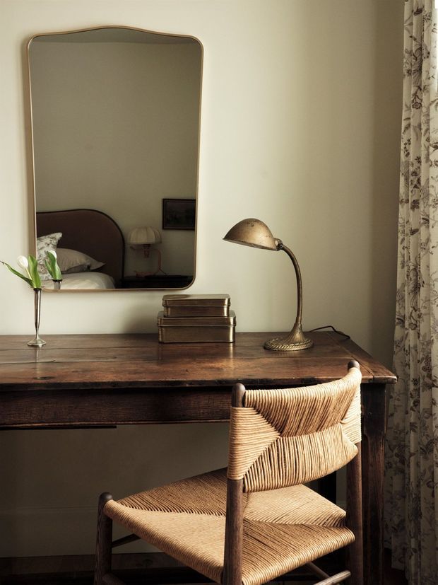 A warm, intimate bedroom workspace or dressing area featuring a dark timber writing desk, woven-seat chair, vintage brass desk lamp, stacked books, and a large rounded rectangular mirror. The mirror reflects part of the bed and bedside table, while patterned curtains frame the right side.