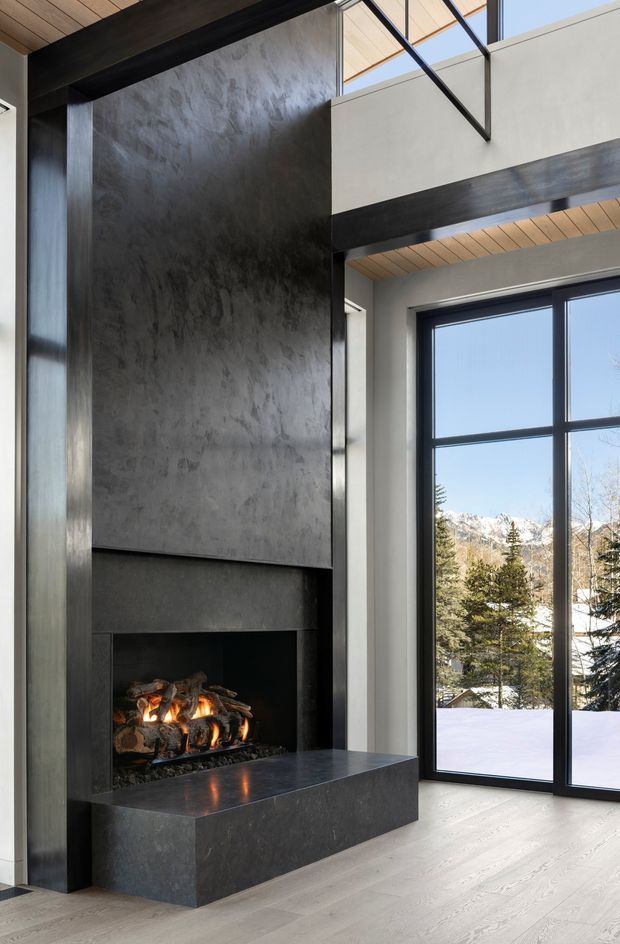 Double-height modern interior featuring a dramatic dark fireplace wall with a broad stone or concrete hearth, exposed black structural beams, light timber ceiling boards, and expansive black-framed glazing. Snow-covered mountain scenery and trees are visible outside.