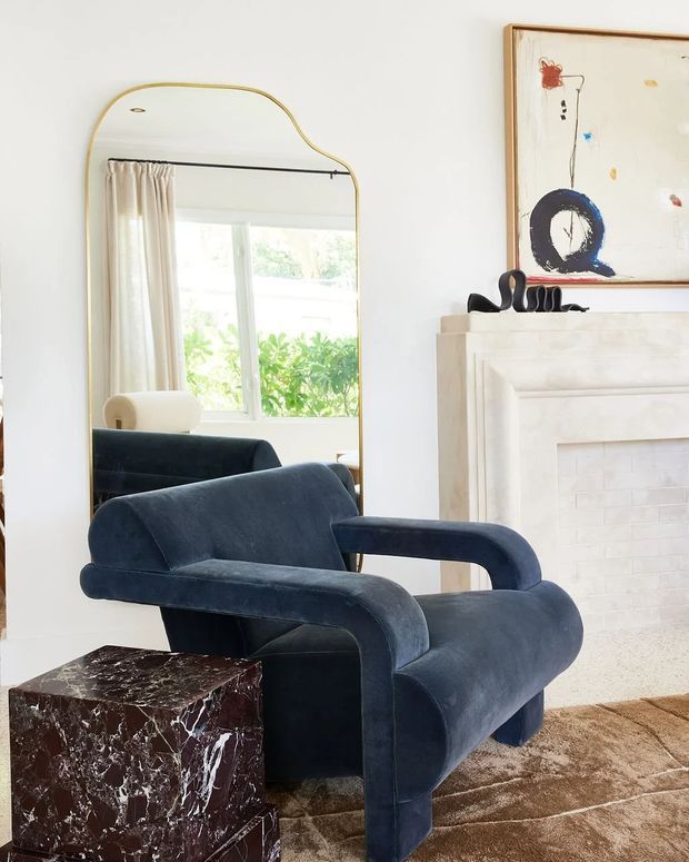 A bright, refined living room vignette featuring a deep navy velvet sculptural armchair beside a dark veined stone side table. A large organic-shaped brass-framed mirror reflects a curtained window and greenery, while a pale painted fireplace surround with white brick infill anchors the room. Abstract artwork and a warm textured rug complete the elegant setting.