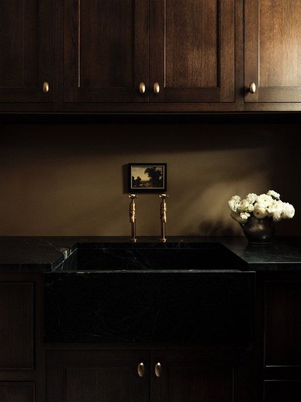 Moody close-up of a richly detailed dark timber kitchen or butler’s pantry, featuring floor-to-ceiling-style upper cabinetry, dark stone benchtops, a large integrated black stone farmhouse sink, brass-toned tapware and cabinet pulls, and warm understated styling with flowers and artwork.