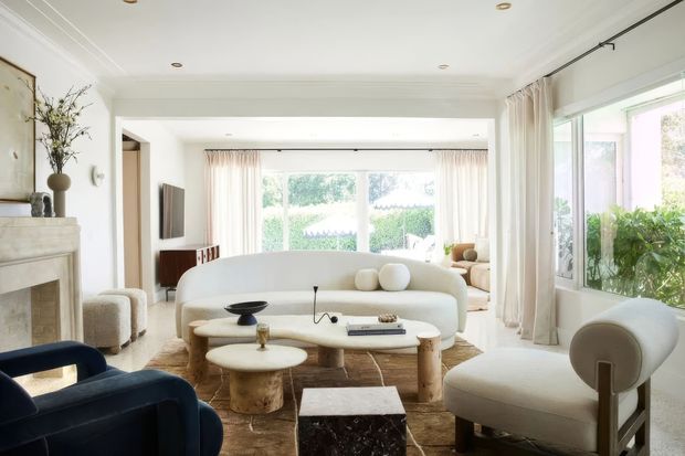 Bright, refined living room with a curved cream sofa, sculptural upholstered armchairs, rounded timber and stone coffee tables, and a dark blue accent chair. The room features a traditional white fireplace, substantial ceiling cornices and beams, large windows with sheer curtains, pale walls, and abundant natural light. A dining or secondary sitting area is visible beyond the main lounge.