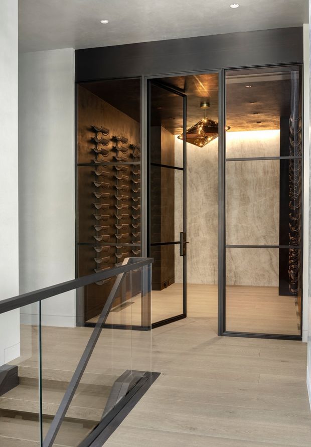 Contemporary wine cellar viewed from a stair landing, enclosed by dark-framed glass doors and partitions. The cellar features extensive wall-mounted wine bottle storage, warm timber-toned surfaces, a textured pale feature wall, light timber flooring, and a sculptural geometric pendant light. A glazed stair balustrade is visible in the foreground.