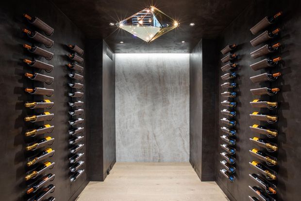 A dark, dramatic wine room with floor-to-ceiling bottle racks mounted along both side walls. The space has charcoal textured walls, a pale stone or concrete feature wall at the rear, light-toned flooring, recessed downlights, and a geometric statement pendant light.