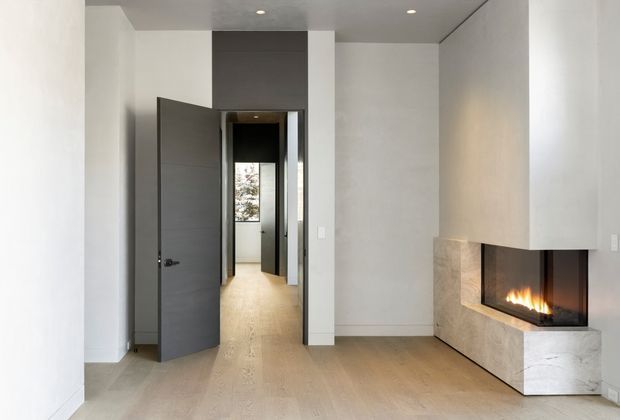 Minimalist contemporary interior showing a quiet living area with pale timber flooring, white walls, a dark grey internal door, recessed ceiling lights, and a built-in corner fireplace clad in light stone. A hallway and additional rooms are visible beyond the open doorway.