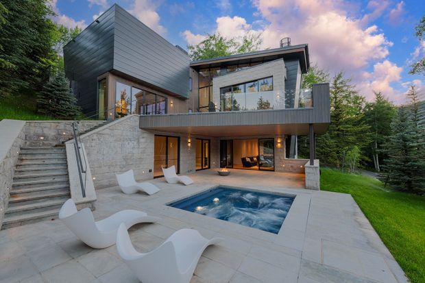 A multi-level contemporary home set into a wooded, sloping site, with dark horizontal cladding, extensive glazing, exposed concrete or pale stone walls, and broad terraces. The foreground features a paved outdoor entertaining area with sculptural white loungers and a small in-ground spa or pool, while an upper balcony projects over the lower level.